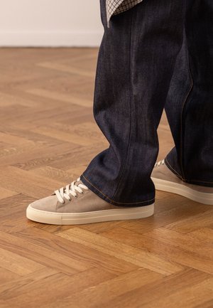Persoon die beige suède sneakers draagt met witte veters en zolen, gecombineerd met donkerblauwe wijde jeans, staand op een houten vloer.