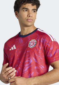 Maillot de football graphique rouge avec des motifs abstraits violets, manches courtes, col bleu marine et bandes blanches Adidas. Présente un blason sur la poitrine.