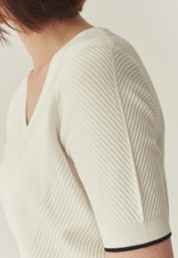 Pull crème en tricot avec col en V, présentant un côtelé texturé et des manches courtes avec une bordure marine au niveau du poignet.