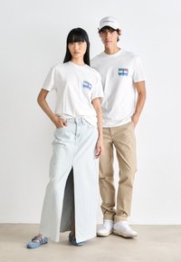 Dos modelos están juntos. La mujer lleva una camiseta blanca con un diseño de bolsillo y una falda de mezclilla azul claro. El hombre viste una camiseta a juego y pantalones beiges.