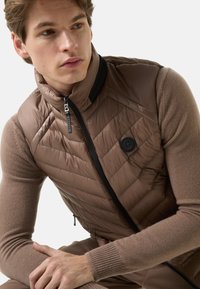 Braune gesteppte Weste mit hohem Kragen, Reißverschluss und Logo-Akzent. Wird über einem hellbraunen Pullover mit gerippten Bündchen getragen. Glatte Textur.