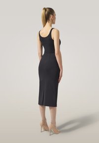 Kvinna står vänd bort, klädd i en svart ärmlös klänning, nude fisknätstrumpor och beige högklackade skor, med håret i en hästsvans.