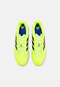Zapatos de fútbol amarillos brillantes, hechos de material sintético, con rayas negras, cuello acolchado y plantillas azules que presentan detalles de marca.