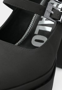 Černá bota Mary Jane se stříbrnou sponou, viditelným šitím a částečně viditelným názvem značky uvnitř.