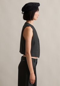 Schwarzes, kurzes Top mit glatter Textur, mit Knopfleiste und weiten Armausschnitten, kombiniert mit hoch taillierten schwarzen Hosen und einer schwarzen Baskenmütze.