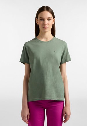 Žena v jednoduchom zelenom tričku s krátkym rukávom a jasne ružových nohaviciach, stojaca pred bielym pozadím, čelom vpred.