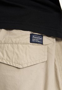 Poche de pantalon beige avec une étiquette Petrol Ind bleu marine indiquant "Denim Driven Since '89 Florida U.S.A" sous l'ourlet d'une chemise noire.