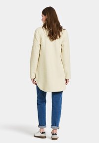 Cremefarvet fleece lang frakke med rundet kant, parret med opsmøgede blå jeans og hvide sneakers med mørke accenter. Ingen synlige lukninger.