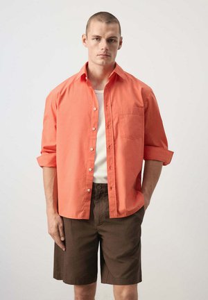 Jeune homme aux cheveux courts portant une chemise corail ouverte à boutons sur un haut blanc et un short marron jusqu’aux genoux, debout devant un fond uni.