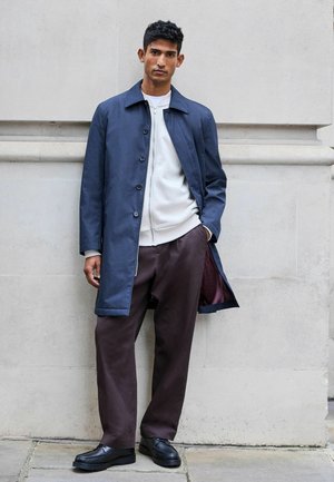 Abrigo impermeable azul marino con cierre de botones, combinado con un suéter gris claro con cremallera y pantalones marrones holgados. Zapatillas negras completan el conjunto.