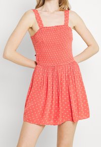 Korálově růžové šaty s nabíraným živůtkem a čtvercovým výstřihem. Mají široká ramínka a rozšířenou sukni s malými bílými květinovými vzory.