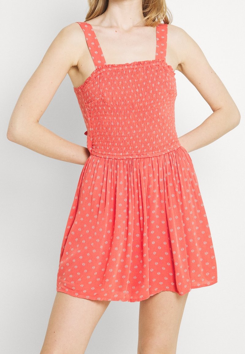 Korálově růžové šaty s nabíraným živůtkem a čtvercovým výstřihem. Mají široká ramínka a rozšířenou sukni s malými bílými květinovými vzory.