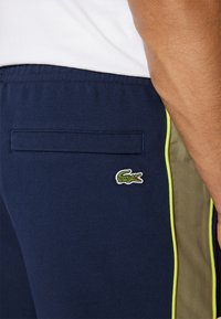 Calças de jogging texturizadas, na cor navy, com detalhes laterais em verde e amarelo, apresentando um bolso traseiro e um pequeno logótipo de crocodilo bordado.