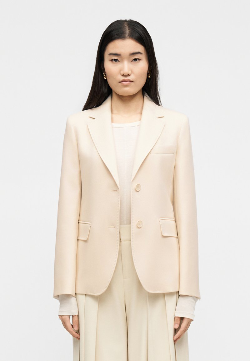 Γυναίκα με μακριά σκούρα μαλλιά που φοράει χρυσούς κρίκους στα αυτιά, κρεμ σακάκι με κουμπιά, ανοιχτόχρωμο μακρυμάνικο τοπ και κρεμ πλισέ παντελόνι μπροστά σε λευκό φόντο.