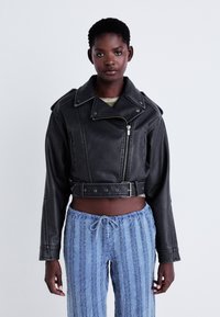Černá zkrácená kožená bunda s páskem v pase, oversized límcem a zapínáním na zip. V kombinaci se světle modrými texturovanými kalhotami.