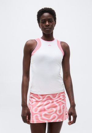 Ženska, ki nosi belo majico brez rokavov z rožnatim robom in rožnato krilo z vzorcem, stoji ob belem ozadju.