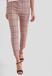 Pantalon à carreaux roses en tissu tissé, avec une coupe ajustée, une longueur raccourcie et un léger éclat, associé à des sandales noires à bride cheville.