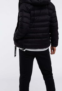 Zwarte donsjack met horizontale stiksels, een capuchon en zijzakken. Draagt over een wit shirt en gecombineerd met zwarte broek.