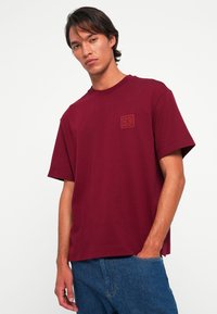Joven vestido con una camiseta suelta de color burdeos con un pequeño logotipo bordado en el pecho y unos pantalones vaqueros azules, con la mano en el bolsillo.