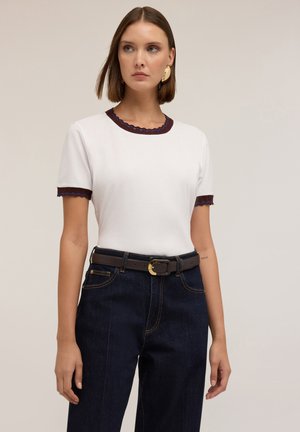 Witte t-shirt met geschulpte donkerrode halslijn en manchetten, gecombineerd met een hoge taille donkere spijkerbroek en een bruine riem met een gouden gesp.