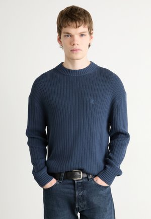 Suéter de punto acanalado azul marino con cuello redondo y mangas largas, usado sobre jeans de mezclilla oscuros, que presenta un sutil logo en el pecho.