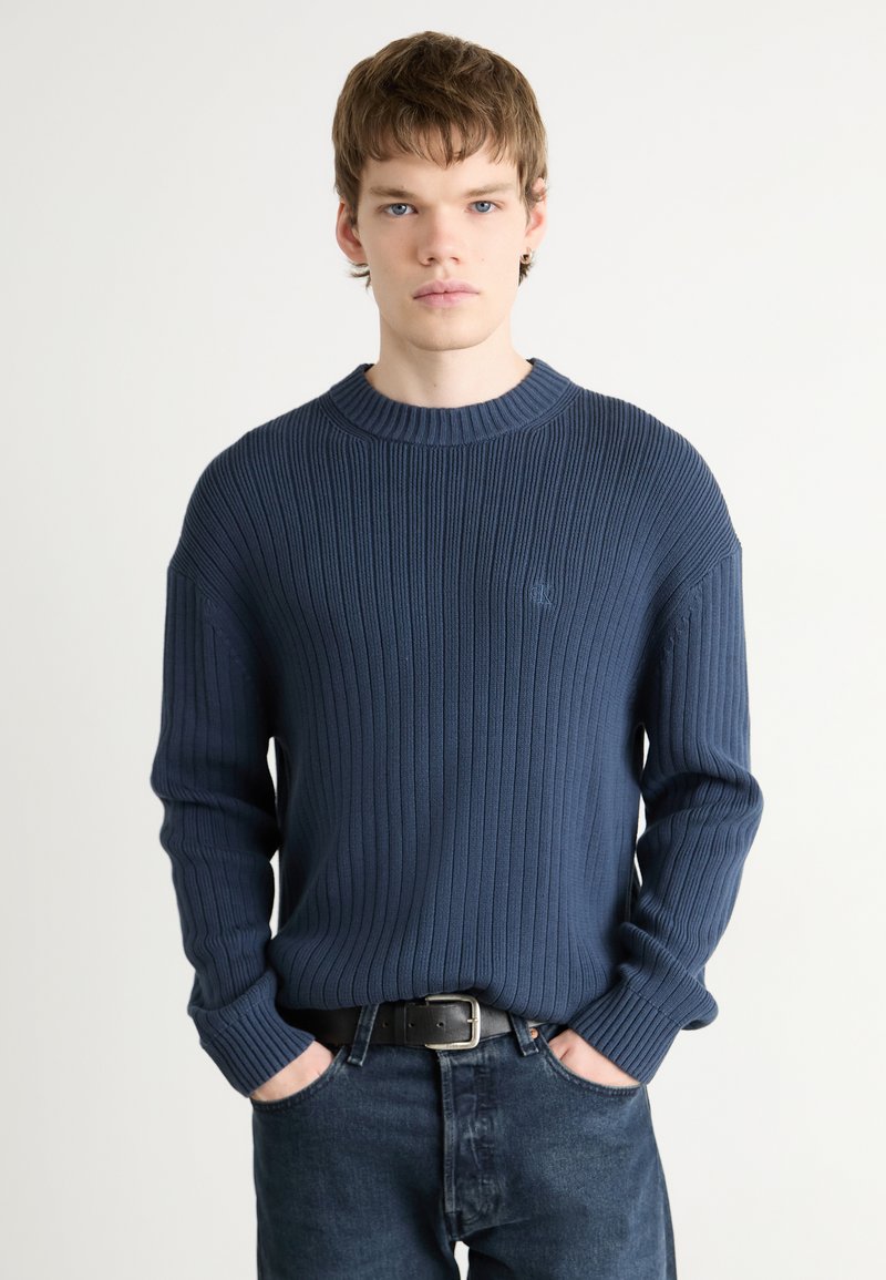 Maglione a coste blu navy con collo tondo e maniche lunghe, indossato su jeans in denim scuri, con un logo discreto sul petto.