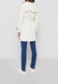 Krémový trenčkot s páskem v pase, ramenními epauletami a knoflíkovými detaily. V kombinaci s modrými džíny a bílými teniskami.