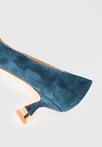 Chaussure pour femme à talon bas en daim bleu, avec un bout pointu et un accent doré sur le talon, sur fond blanc.