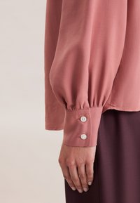 Blusa a maniche lunghe in rosa polveroso, con polsini plissettati e due bottoni bianchi. Tessuto morbido, vestibilità rilassata, abbinata a un capo di colore viola scuro.