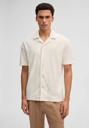 Mężczyzna stoi zwrócony przodem, ubrany w białą teksturowaną koszulę na krótki rękaw z guzikami oraz jasnobrązowe spodnie, na jednolitym szarym tle.