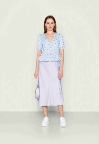 Top peplum azul claro com estampa floral, combinado com uma saia lilás. Footwear são ténis brancos. A modelo segura uma pequena bolsa lavanda.
