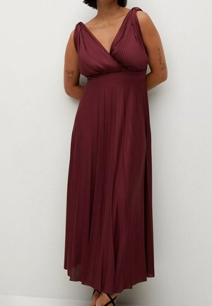 Kvinde iført ærmeløs, dyb bordeaux plisseret maxikjole med V-hals og skulderknuder, stående mod en hvid væg med armene bag ryggen.