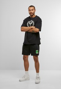 Man stands with arms crossed wearing black graphic T-shirt, black shorts with yellow-green emblem, white socks, and white sneakers on plain background.
