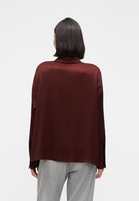 Oversized bordeauxbluse i letvægtsstof med krave, der har en diskret glans og nedsænkede skuldre, stylet med grå bukser.