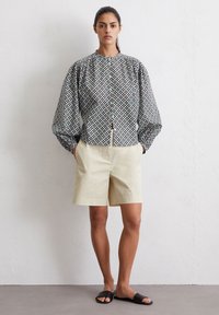 Langarmhemd mit geometrischem Druck in Blau, Schwarz und Weiß; verfügt über puffärmel. Kombiniert mit beigen Shorts und schwarzen Sandalen.