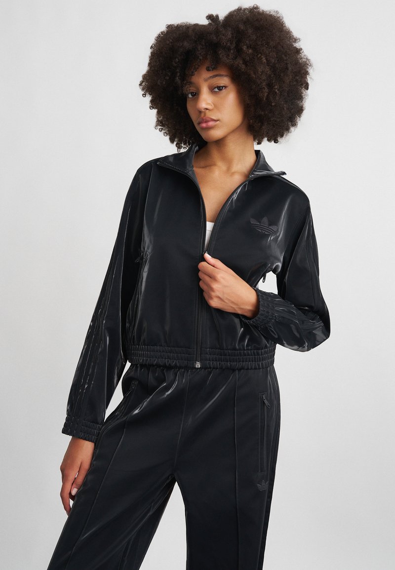 Juodas poliesterinis sportinis kostiumėlis su stovinčia apykakle, užtrauktuku priekyje ir elastingomis rankovėmis. Yra toninių trijų juostų raštas ir logotipas.