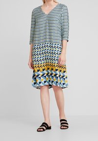 Vestido estampado con escote en V, que presenta diseños geométricos en azul, amarillo y negro. Tejido suave y mangas de tres cuartos. Sandalias de tacón bajo negras.
