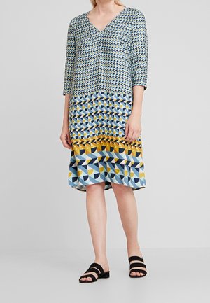 Robe à motifs avec un décolleté en V, présentant des designs géométriques en bleu, jaune et noir. Tissu doux et manches trois-quarts. Sandales noires à enfiler.