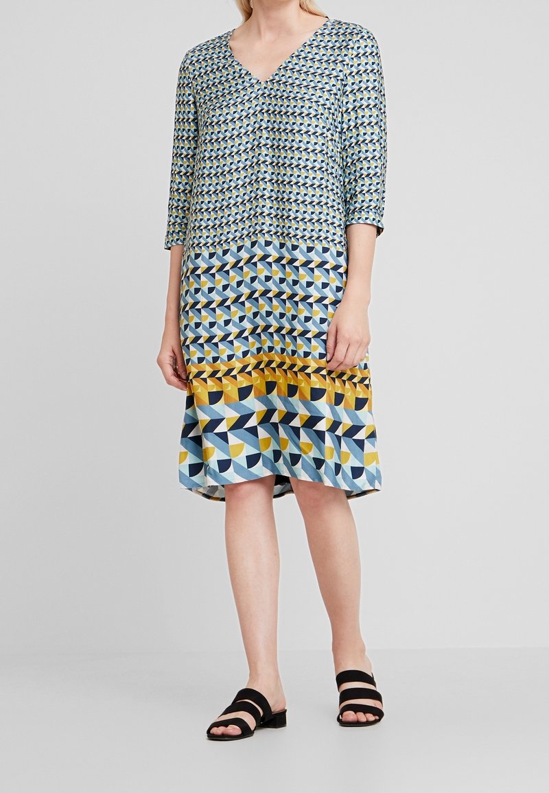 Vestido estampado con escote en V, que presenta diseños geométricos en azul, amarillo y negro. Tejido suave y mangas de tres cuartos. Sandalias de tacón bajo negras.
