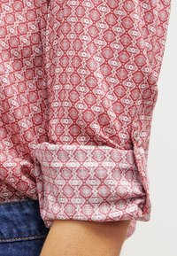 Chemise à motifs rouges avec des dessins géométriques blancs, présentant un poignet boutonné et un tissu texturé, partiellement visible sur un fond en denim.