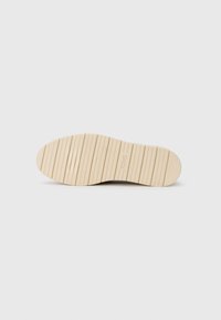 Suela de zapato de goma beige con ranuras verticales y bordes redondeados, con un sutil logotipo en el centro. Textura suave en general.