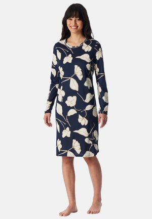 Smiling woman stands barefoot wearing a navy knee-length dress with long sleeves and large white floral print.