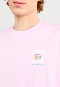 Jasnoróżowa bawełniana koszulka z okrągłym dekoltem, z wielokolorowym graficznym tekstem "MŁODZIEŻ ZJEDNOCZONA" i "SĄ PRZYSZŁOŚCIĄ" w żywej typografii.