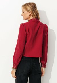Blouse rouge texturée avec des manches bouffantes et des dentelles au col et aux poignets, portée avec un jean foncé, vue de dos.