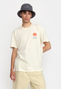 Camiseta de algodón crema con mangas cortas, que presenta un gráfico circular naranja y el texto "Revolución". Llevada con pantalones cortos beige y un sombrero de cubo gris.