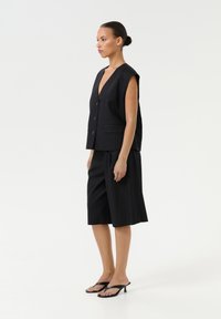 Črna brezrokavnik s V-izrezom, tremi gumbi in dvema sprednjima žepoma, v kombinaciji s črnimi kratkimi hlačami do kolen in črnimi sandali s peto.