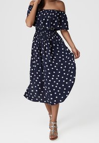 Námorně modré midi šaty bez ramínek s bílými puntíky, nošené se stříbrnými sandály na svůdné pásky, modelka drží lem sukně.