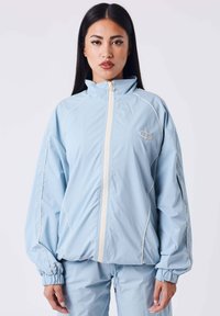 Giacca a vento azzurro chiaro con colletto alto, dettagli bianchi e polsini elasticizzati. Presenta una zip frontale e un design con vestibilità ampia.