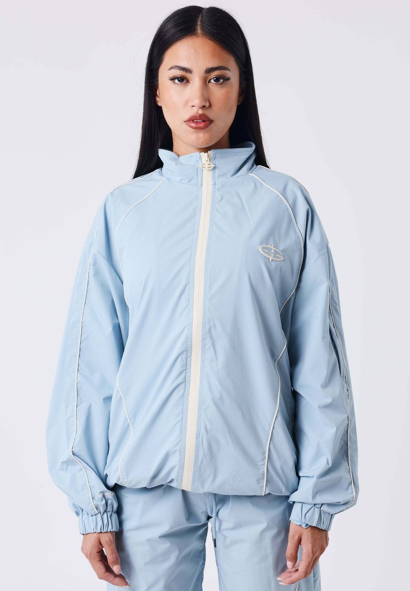 Giacca a vento azzurro chiaro con colletto alto, dettagli bianchi e polsini elasticizzati. Presenta una zip frontale e un design con vestibilità ampia.