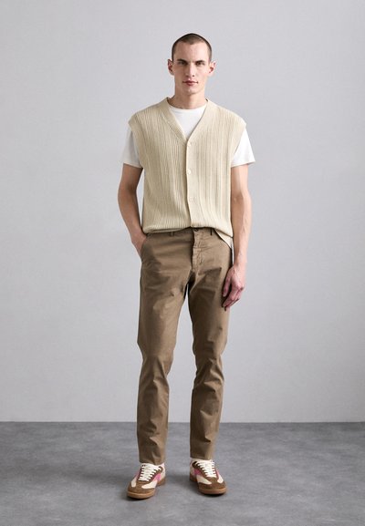 Kreemivärvi ribakootud vest valge T-särgi peal, kombineeritud khaki pükstega ja pruunide spordijalatsitega, millel on roosa aktsent. Lihtne, minimalistlik esteetika.